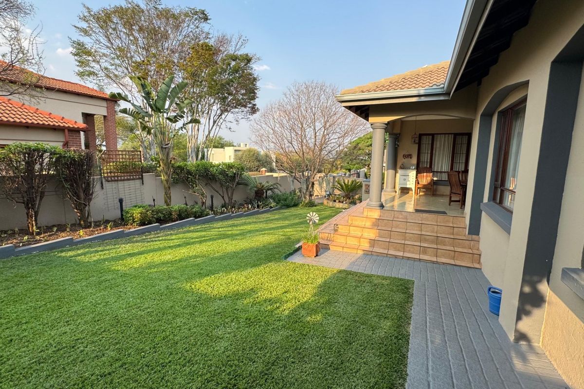 A well-maintained rear garden featuring a tiled patio with concrete columns, a paved walkway, and a neatly manicured lawn enclosed by a boundary wall.