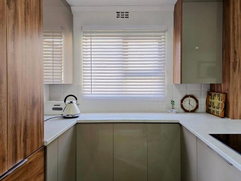 Modern kitchen featuring sleek high-gloss cabinetry, wood-grain accents, white laminate countertops, and a tiled backsplash with integrated hob and venetian blinds.