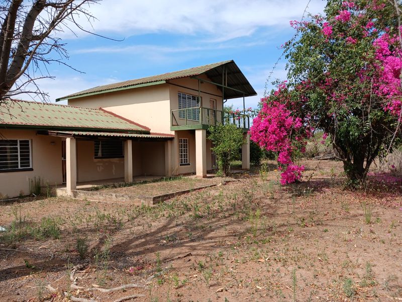 A two-story farm house exterior featuring a covered patio, upper balcony, and metal roofing, set within a natural, unlandscaped garden environment.