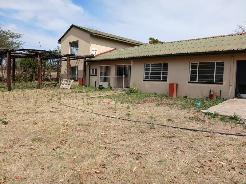 Rear view of the farm property featuring a metal roof, security gates, and a timber pergola structure set within an expansive, unmaintained garden area.