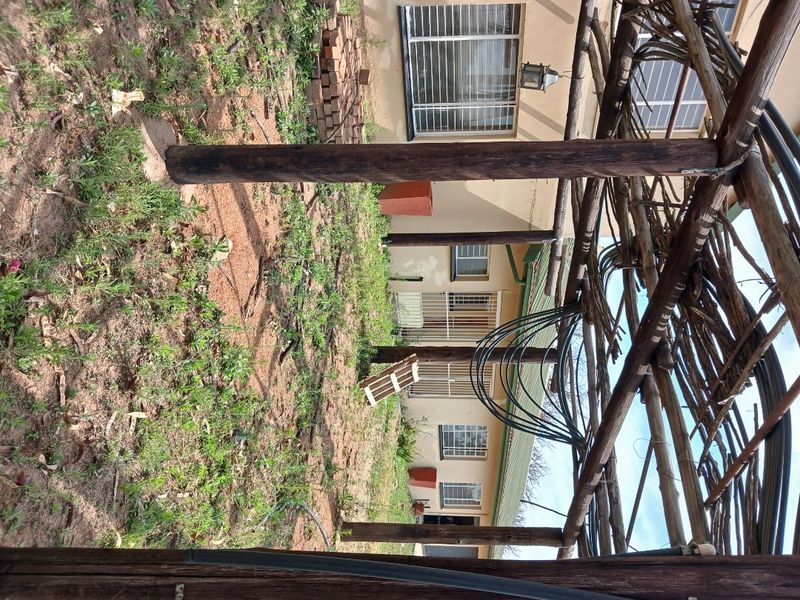 Rear exterior view of the property featuring a weathered timber pergola structure, security-fitted windows, and an unlandscaped garden area requiring maintenance.