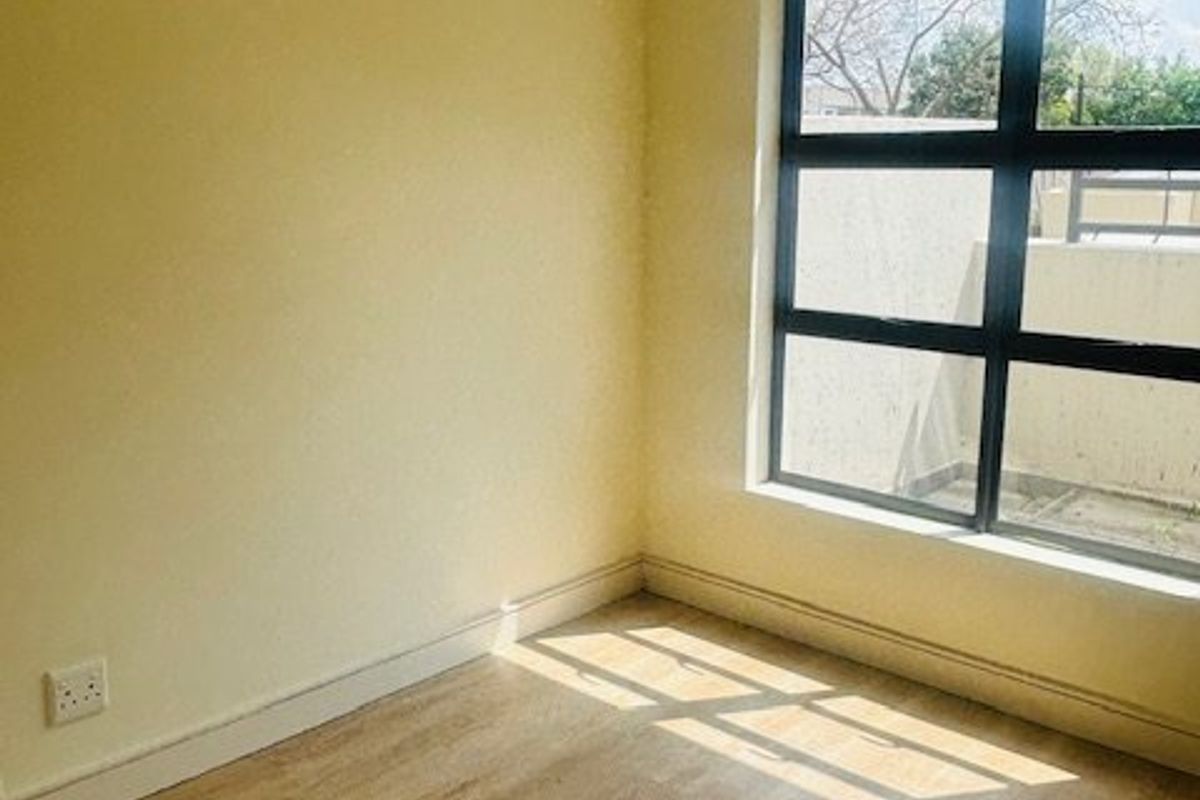 A bright, empty bedroom featuring light-toned laminate flooring, neutral walls, and a large aluminium-framed window allowing for natural light.
