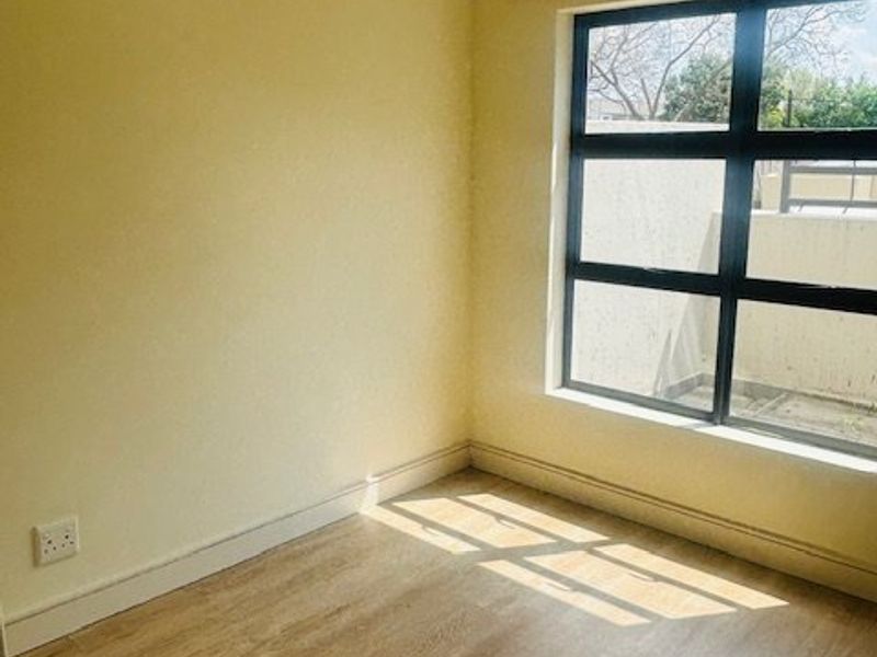 A bright, empty bedroom featuring light-toned laminate flooring, neutral walls, and a large aluminium-framed window allowing for natural light.