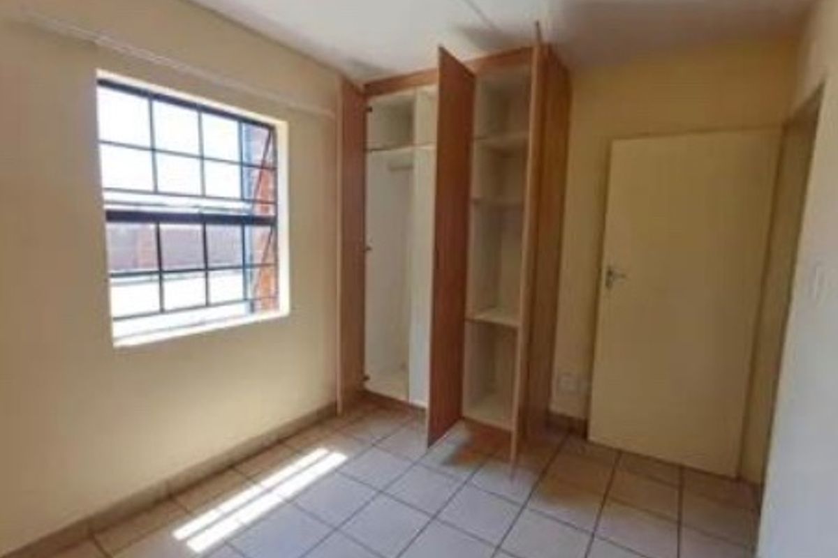 A simple, empty bedroom featuring tiled flooring, a grid-pattern window, and built-in wooden cupboards.