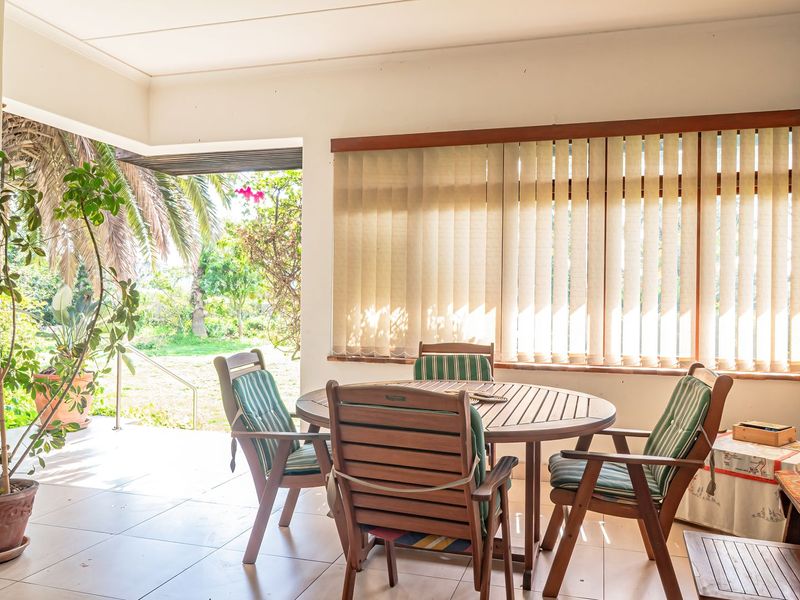 A covered patio area featuring tiled flooring and large windows with vertical blinds, offering direct access to the garden.