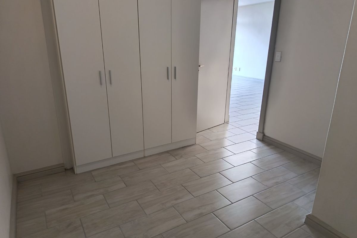 A bright, empty bedroom featuring neutral wood-look tiled flooring and ample built-in cupboard storage, leading into an adjacent room.