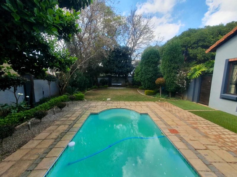 A well-maintained backyard featuring a swimming pool with stone paving, surrounded by established trees and a landscaped garden area.