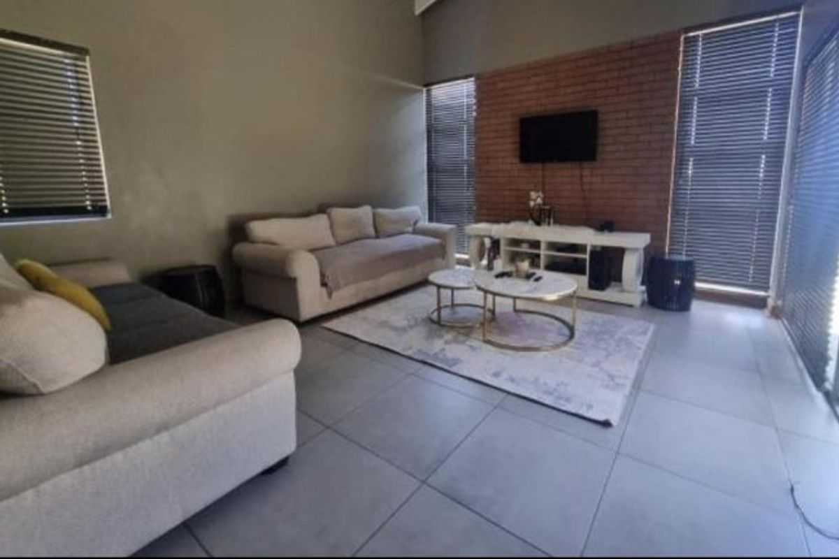 Spacious living room featuring a striking exposed brick feature wall, large format floor tiles, and vaulted ceilings with natural light through venetian blinds.
