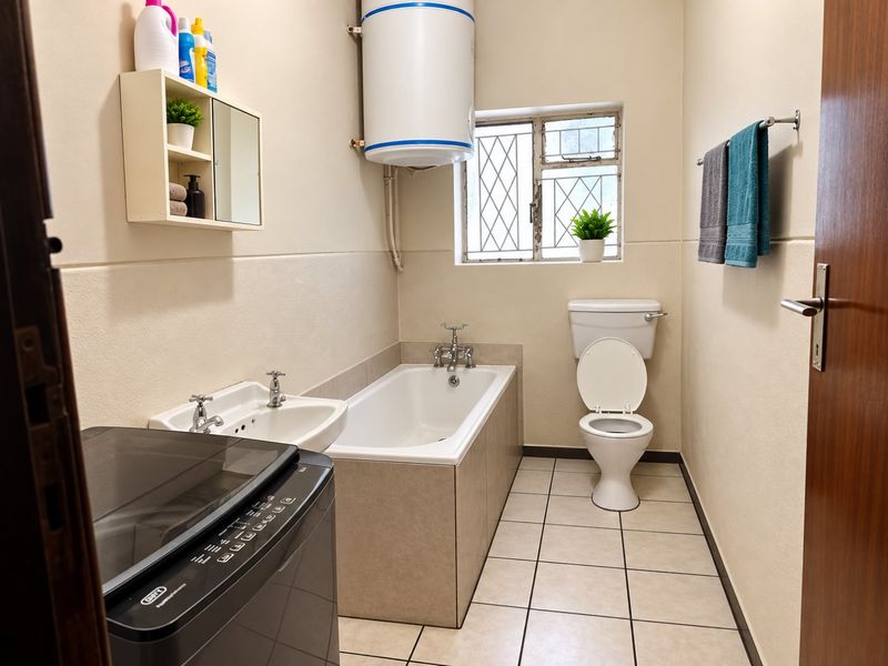 A functional bathroom featuring a bathtub, pedestal basin, toilet, and an electric water heater, with tiled flooring and security bars on the window.