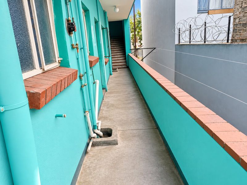 An exterior apartment balcony walkway featuring a brick-topped parapet wall, concrete flooring, and exposed external plumbing infrastructure.