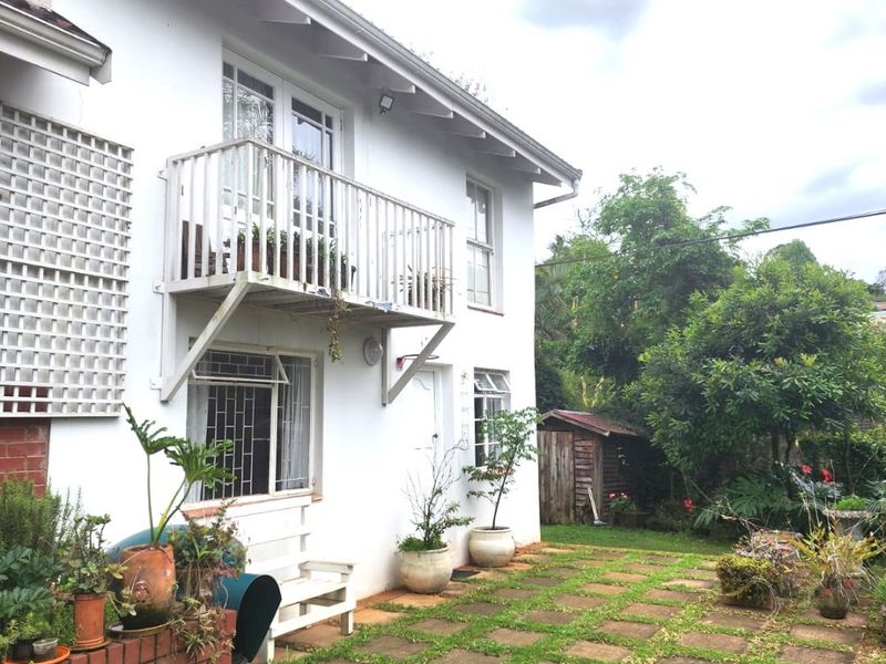 Rear exterior view of the apartment featuring a balcony, paved patio area with brick retaining wall, and a small garden with a storage shed.
