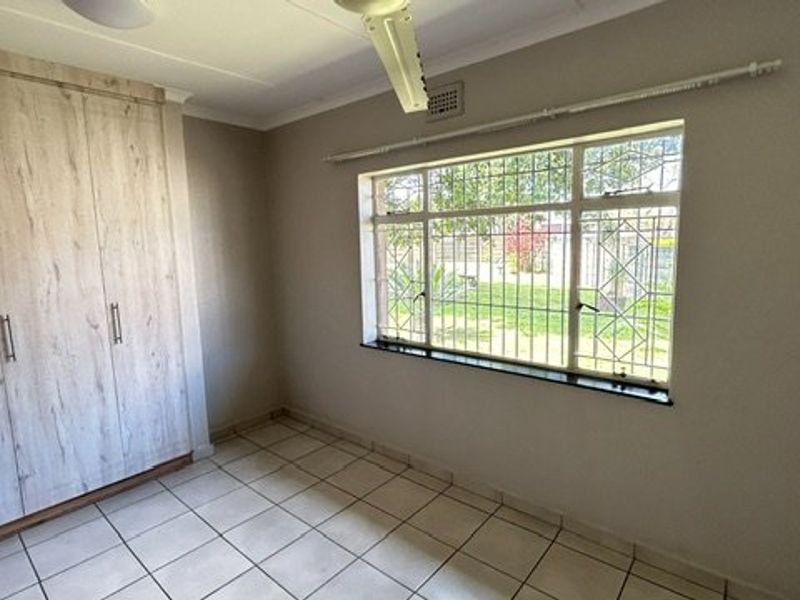 A bright, empty bedroom featuring tiled flooring, built-in cupboards, a ceiling fan, and large windows with security bars.