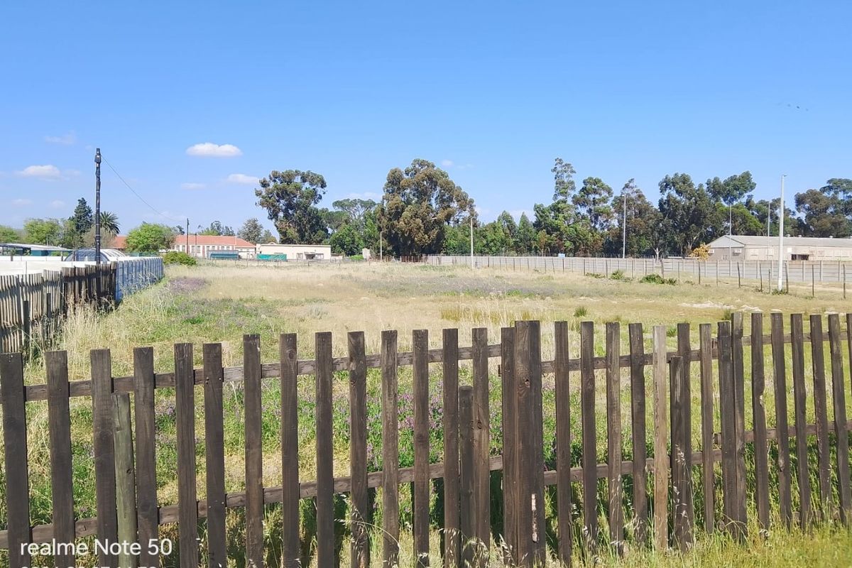 A view of a vacant residential plot enclosed by wooden and wire fencing, featuring open grass and surrounding trees.