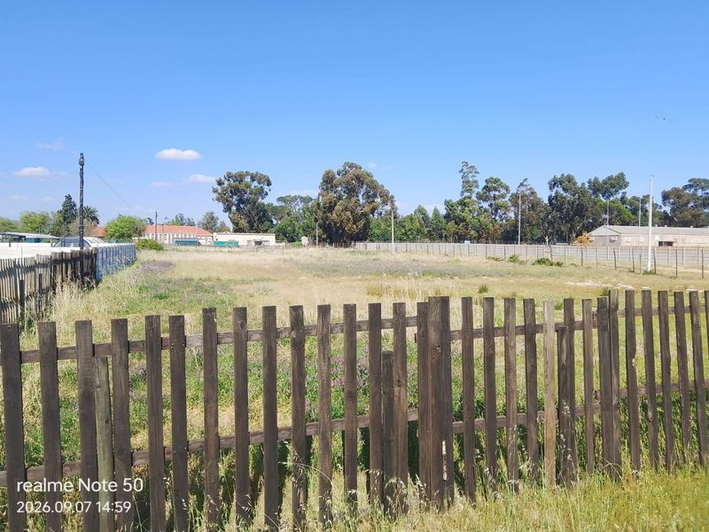 A view of a vacant residential plot enclosed by wooden and wire fencing, featuring open grass and surrounding trees.
