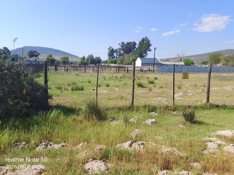 A vacant residential plot featuring natural grass coverage and perimeter wire mesh fencing with wooden posts.