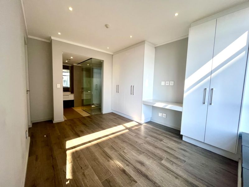 A bright, modern bedroom featuring built-in cupboards, a integrated desk, laminate flooring, and an en-suite bathroom with a glass shower enclosure.