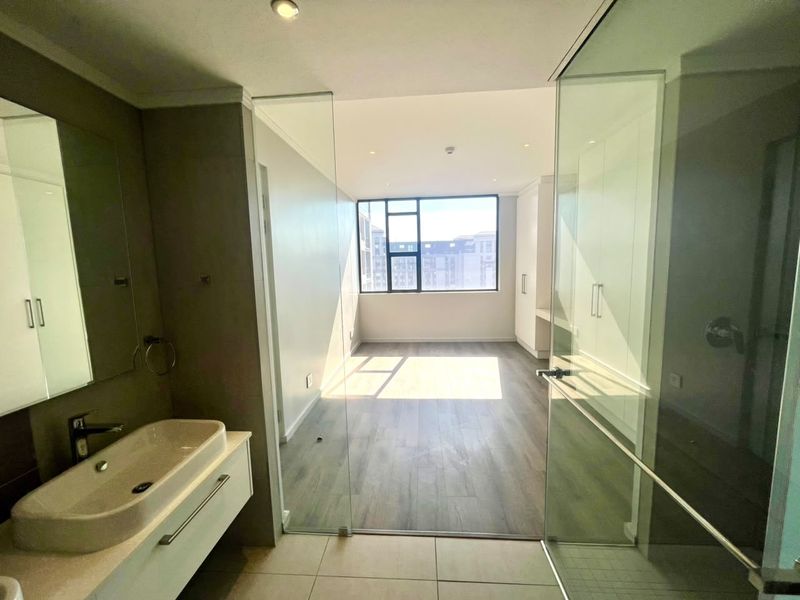 A bright, modern bedroom featuring laminate flooring, built-in cupboards, and an en-suite bathroom with a glass-enclosed shower and vanity.