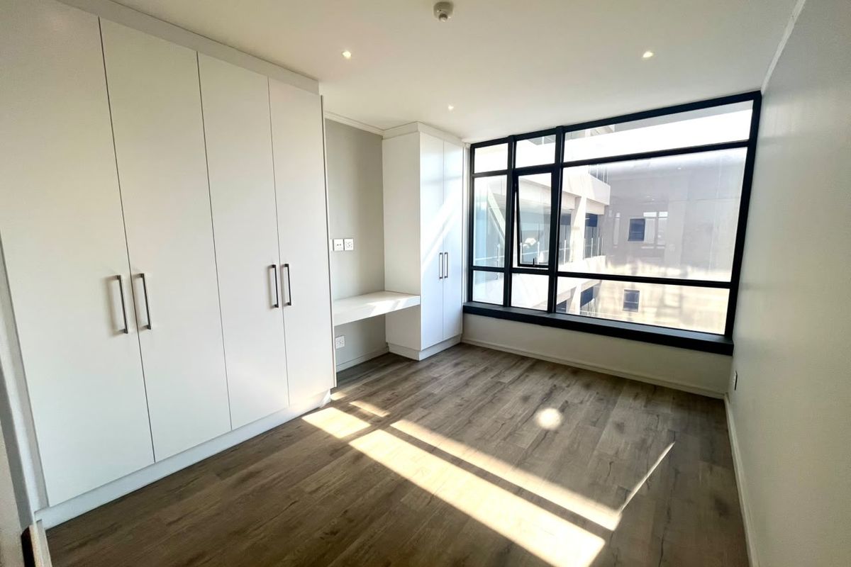 A bright, modern bedroom featuring sleek built-in cupboards, a integrated desk, laminate flooring, and large windows providing ample natural light.