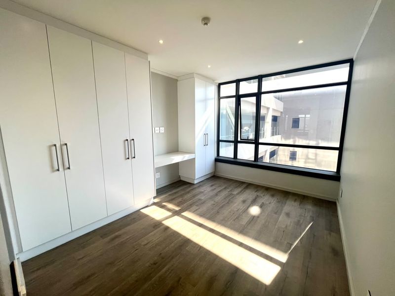 A bright, modern bedroom featuring sleek built-in cupboards, a integrated desk, laminate flooring, and large windows providing ample natural light.