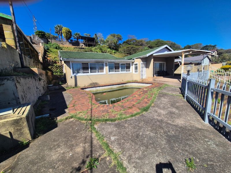 Rear exterior view of the house featuring a swimming pool, concrete patio area, and a carport, set against a sloped garden backdrop.
