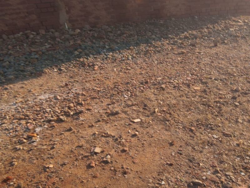 A view of a brick exterior wall with a security window grille, showing uneven ground and debris in the yard area.