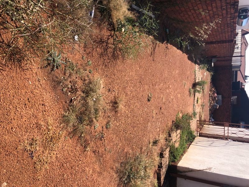 A vacant residential plot area featuring a dirt ground surface, enclosed by brick boundary walls and a metal gate.