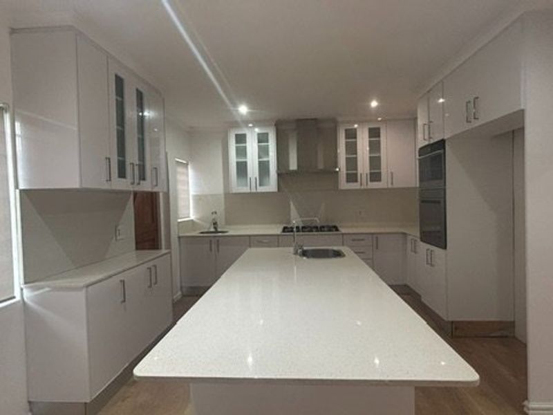 Modern kitchen featuring a large central island with quartz countertops, built-in oven, gas hob, and ample white cabinetry with glass-fronted display units.