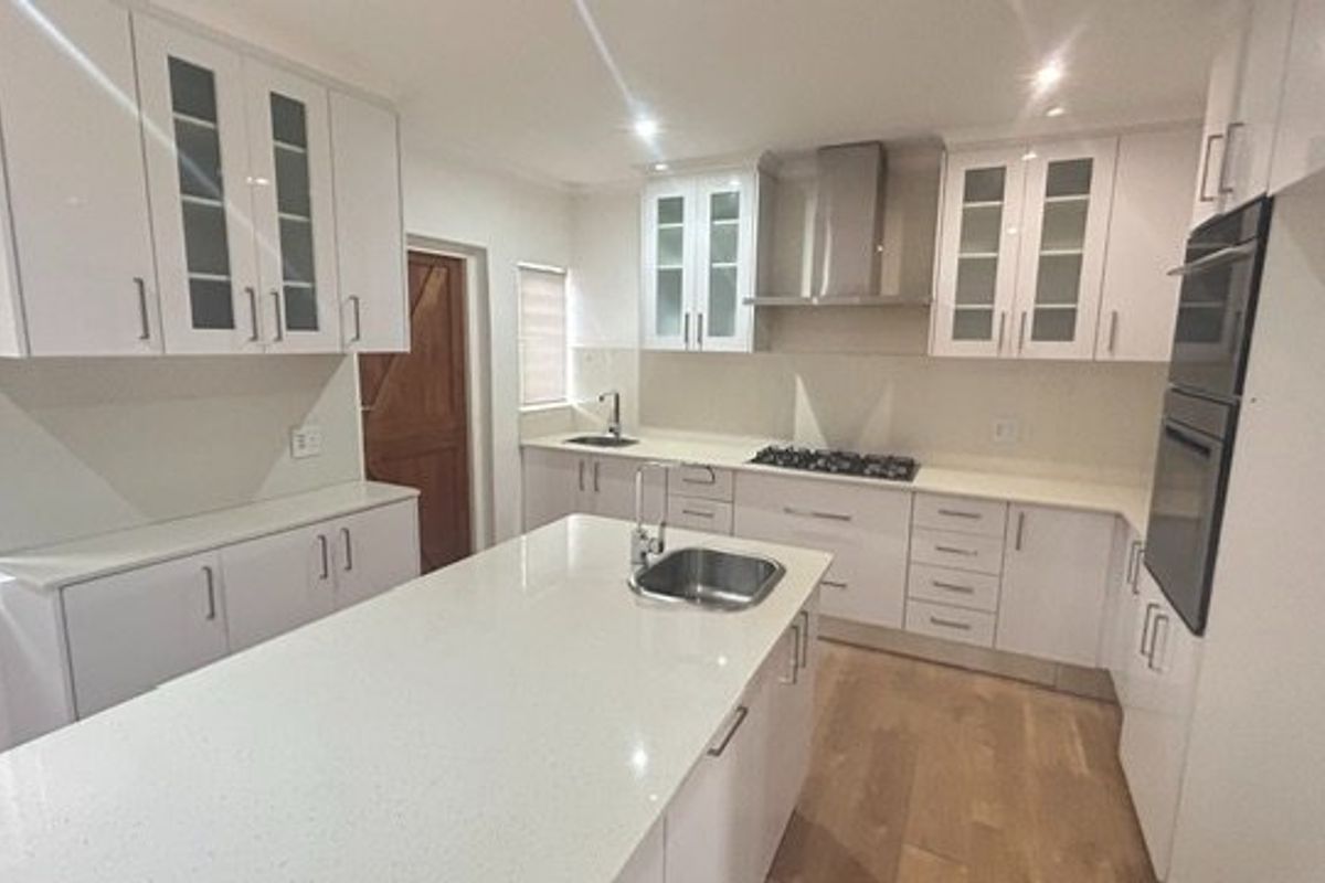 Modern kitchen featuring white cabinetry, stone countertops, a central island with sink, gas hob, and built-in oven, complemented by timber flooring.