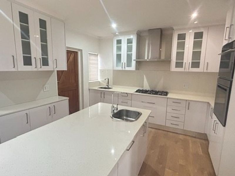 Modern kitchen featuring white cabinetry, stone countertops, a central island with sink, gas hob, and built-in oven, complemented by timber flooring.