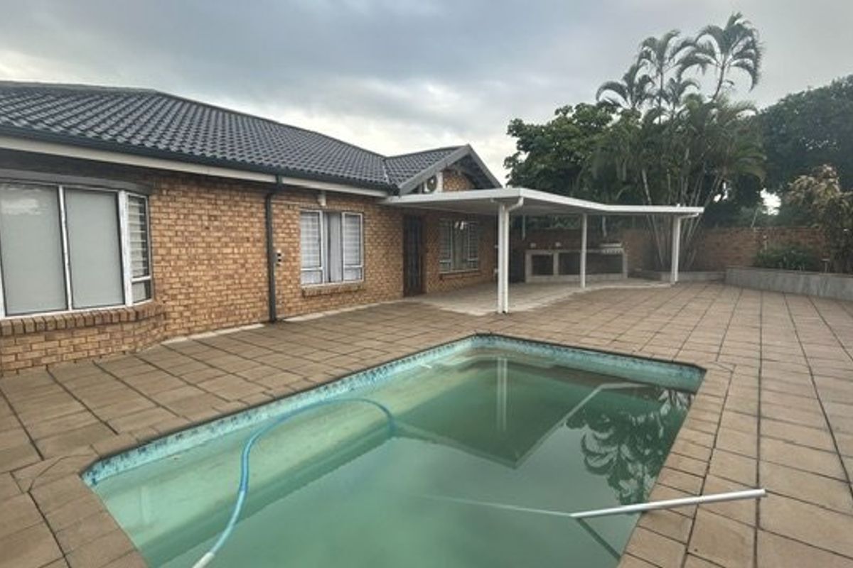 Rear exterior view featuring a brick facade, a covered patio with a built-in braai, and a swimming pool set within a paved outdoor area.