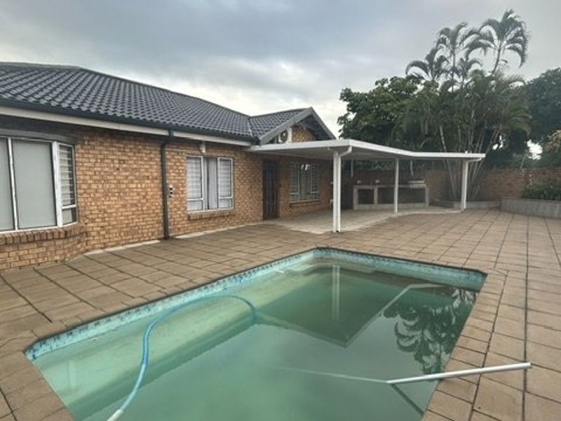 Rear exterior view featuring a brick facade, a covered patio with a built-in braai, and a swimming pool set within a paved outdoor area.