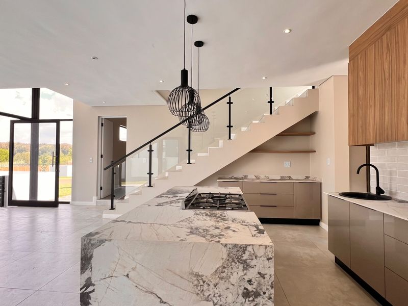 Modern open-plan kitchen featuring a large stone-topped island, gas hob, sleek cabinetry, and a striking staircase with glass balustrade in a bright, contemporary space.