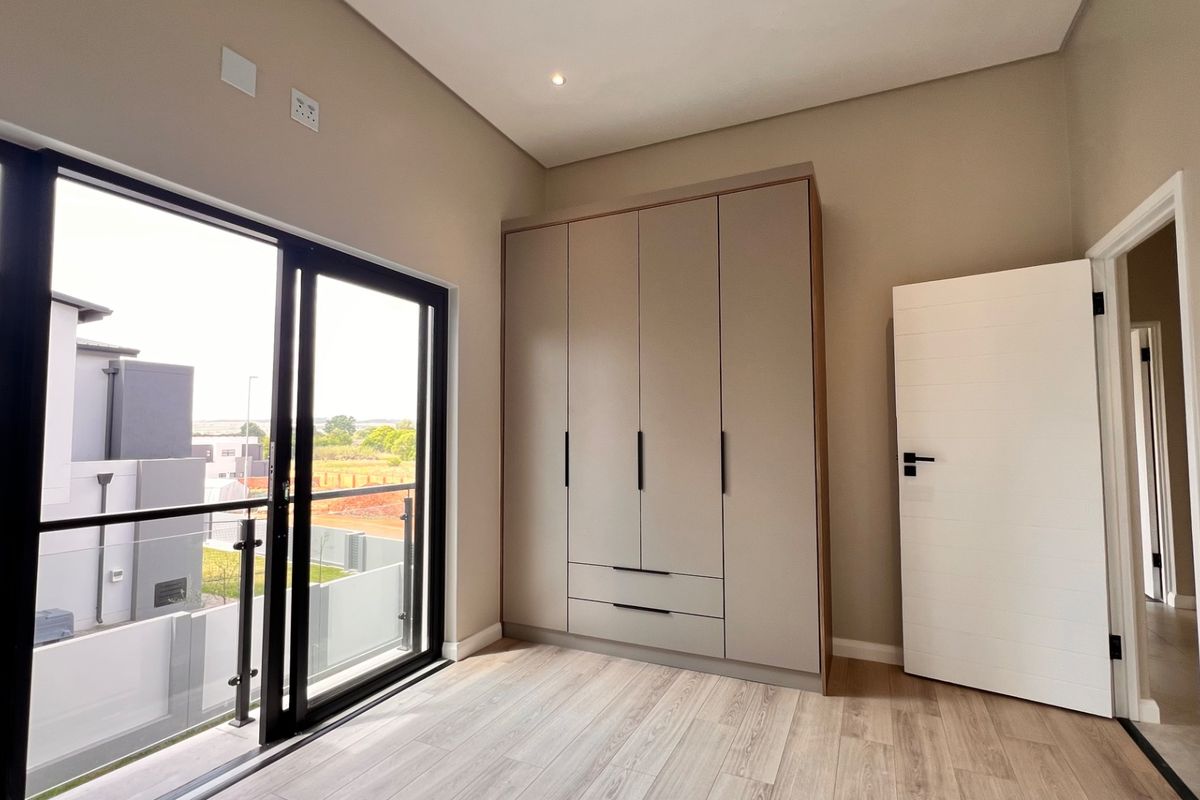 A bright, modern bedroom featuring neutral tones, built-in cabinetry, laminate flooring, and a sliding glass door leading to a private balcony.