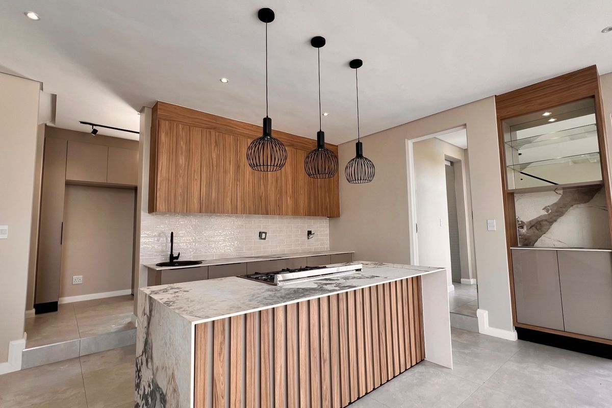 Modern kitchen featuring a large stone-topped island with built-in hob, sleek wooden cabinetry, integrated display shelving, and contemporary pendant lighting.