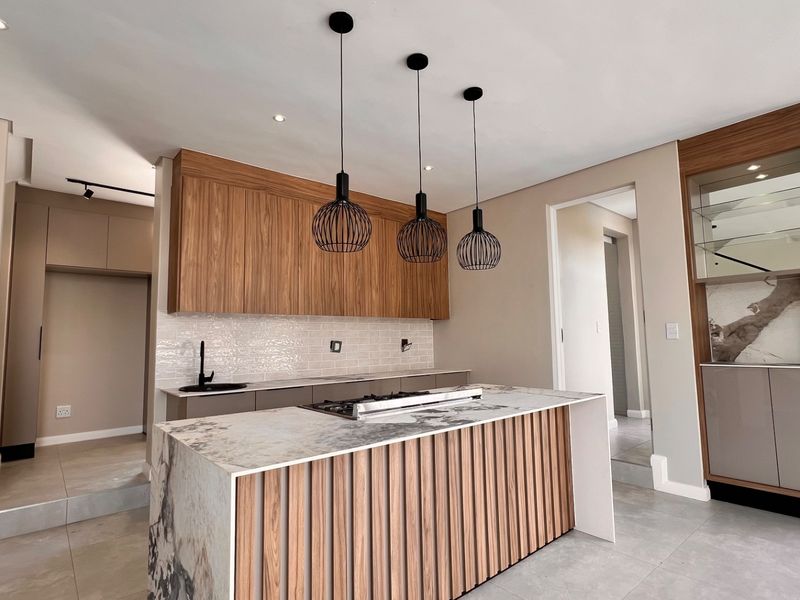 Modern kitchen featuring a large stone-topped island with built-in hob, sleek wooden cabinetry, integrated display shelving, and contemporary pendant lighting.