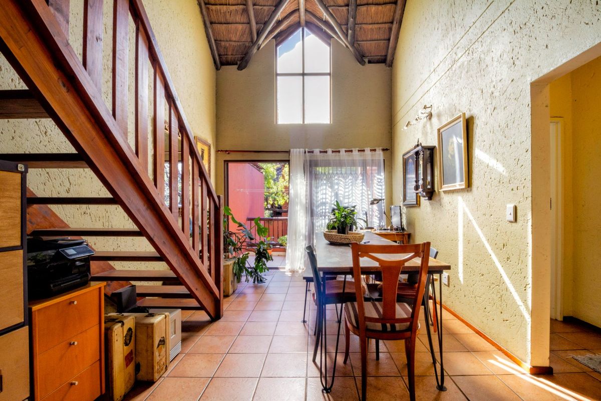 Open-plan living area featuring a double-volume thatched ceiling, exposed timber rafters, a wooden staircase, and ceramic tiled flooring with access to a patio.