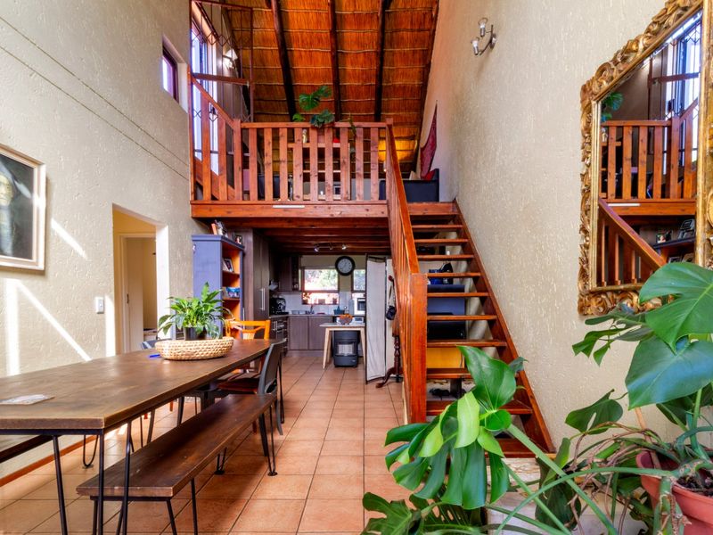 Open-plan living area featuring a double-volume thatched ceiling, exposed timber rafters, a mezzanine level with timber staircase, and tiled flooring throughout.