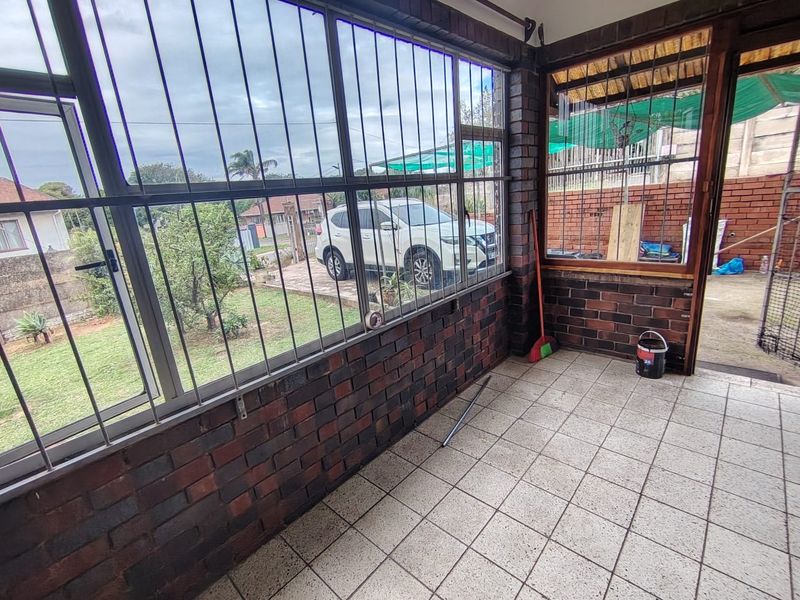 A covered patio area featuring exposed brick walls, tiled flooring, and large windows with security bars, offering a view of the front garden.