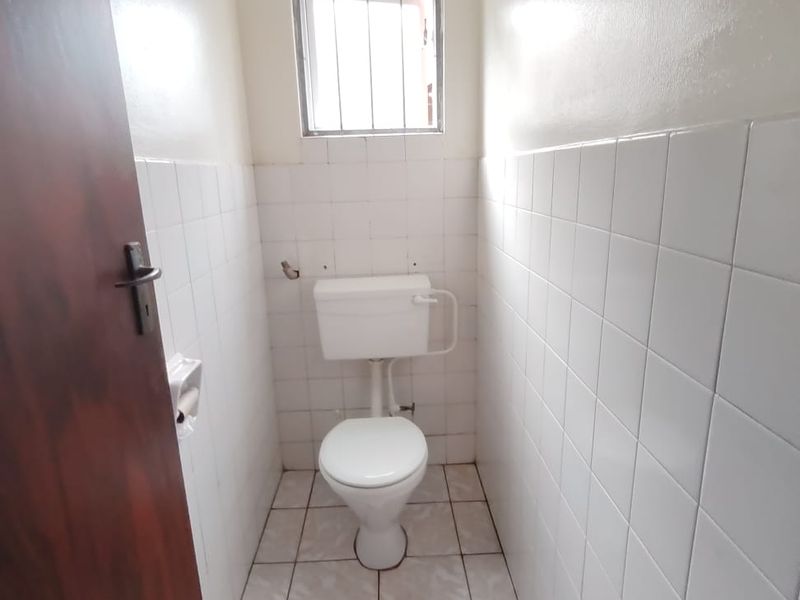 A compact, functional guest bathroom featuring white ceramic wall and floor tiles, a standard toilet, and a small window with security bars.
