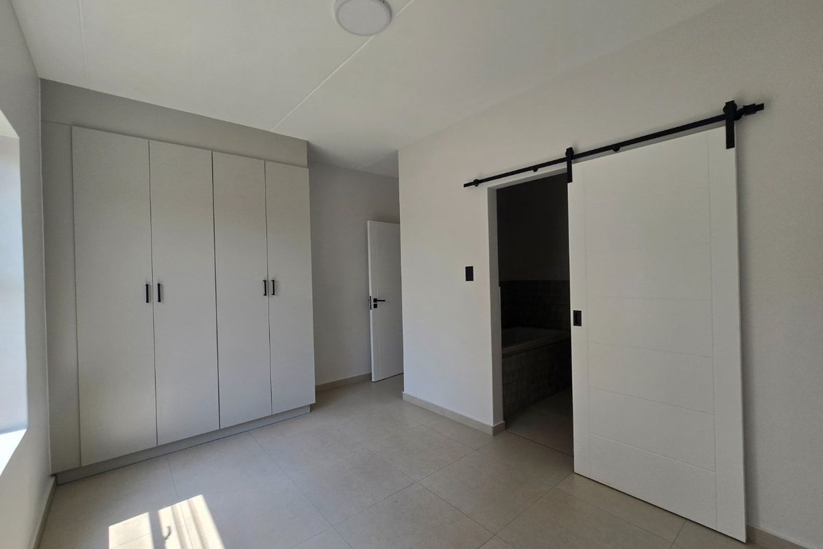 A bright, modern bedroom featuring neutral tiled flooring, ample built-in cupboard storage, and a contemporary sliding barn door leading to an en-suite bathroom.