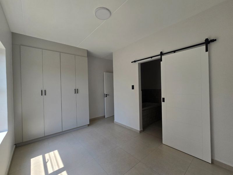 A bright, modern bedroom featuring neutral tiled flooring, ample built-in cupboard storage, and a contemporary sliding barn door leading to an en-suite bathroom.