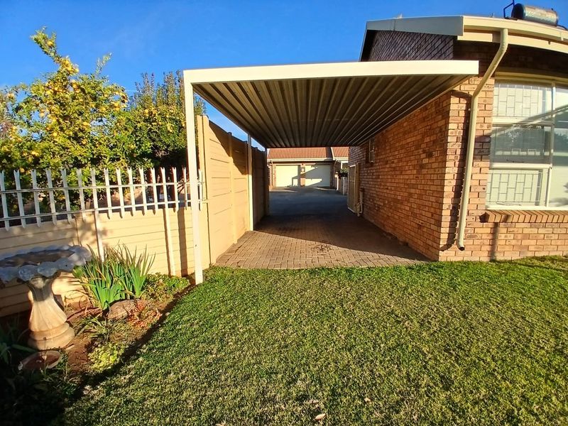A well-maintained townhouse exterior featuring a covered carport, paved driveway, brick facade, and a neat front garden area.