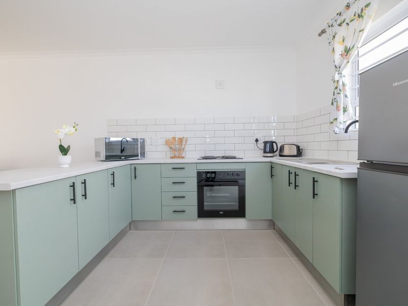 A modern, U-shaped kitchen featuring sage green cabinetry, white subway tile backsplash, built-in oven, electric hob, and neutral tiled flooring.