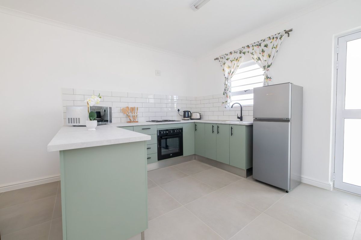 A clean, modern kitchen featuring sage green cabinetry, subway tile backsplash, built-in oven, hob, and neutral tiled flooring with direct exterior access.