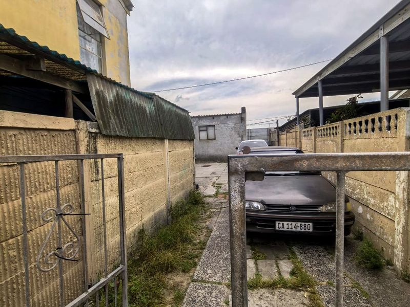 A narrow driveway entrance featuring a metal gate, precast concrete boundary walls, and a carport, showing significant signs of weathering and maintenance requirements.