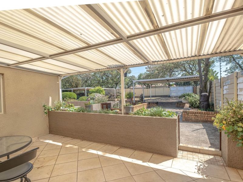 Covered patio area featuring tiled flooring, a built-in brick planter box, and a corrugated roof, overlooking a paved rear garden space.