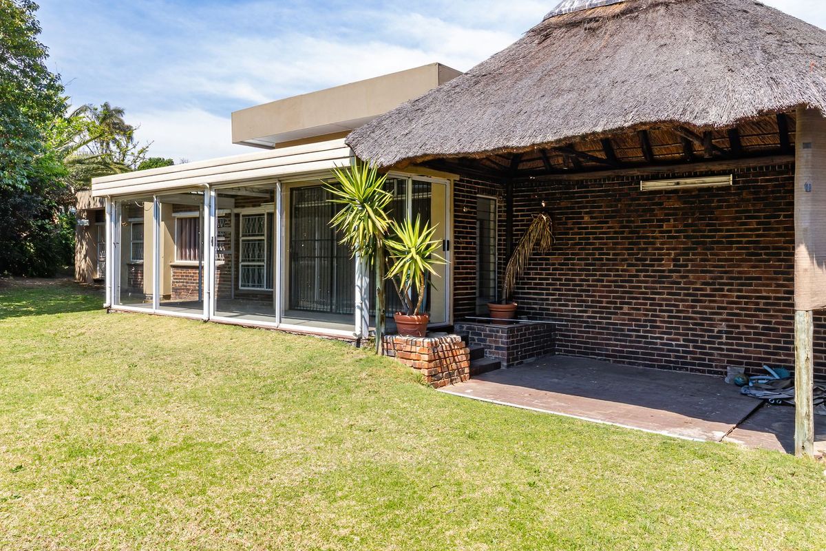 Rear view of the property featuring a spacious lawn, an enclosed patio area, and a traditional thatched roof lapa with exposed brick walls.