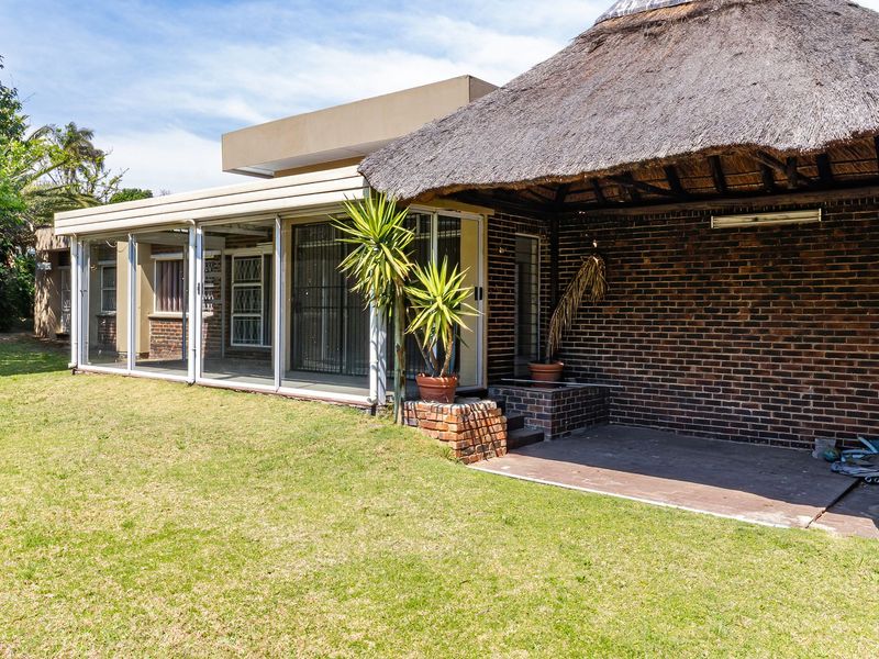 Rear view of the property featuring a spacious lawn, an enclosed patio area, and a traditional thatched roof lapa with exposed brick walls.
