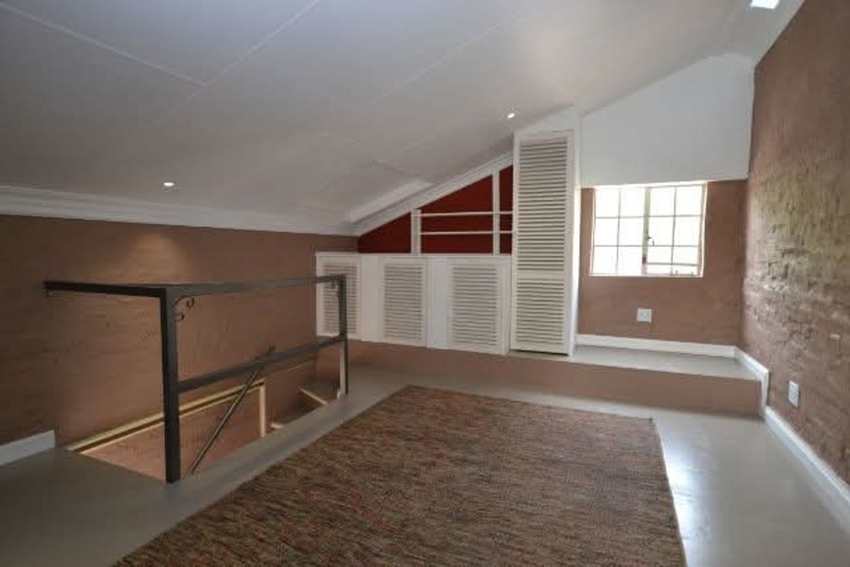 A spacious loft-style bedroom featuring a sloped ceiling, exposed brick wall, built-in storage cupboards, and a metal staircase railing.