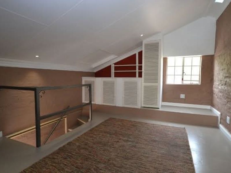 A spacious loft-style bedroom featuring a sloped ceiling, exposed brick wall, built-in storage cupboards, and a metal staircase railing.